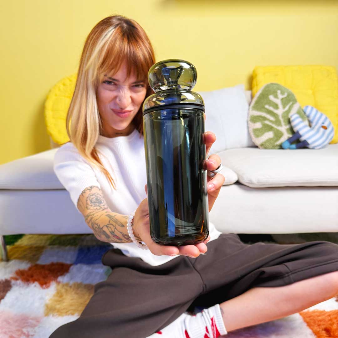 model sitting on colorful checkered rug with white couch in the background. Model is holding up a black glass gravity bong. 