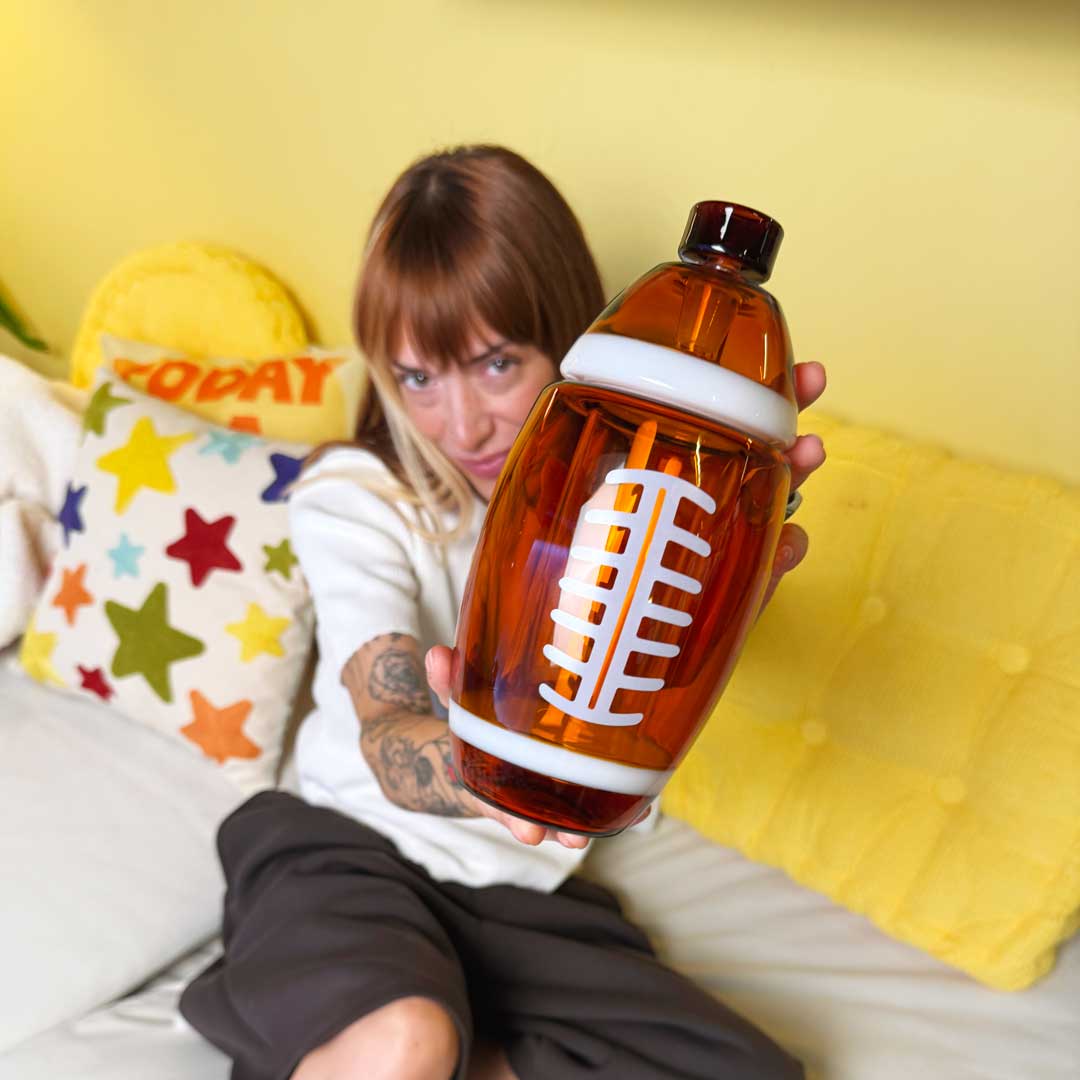 model sitting on white couch with colorful throw pillows. Model is showing off an American football gravity bong. 