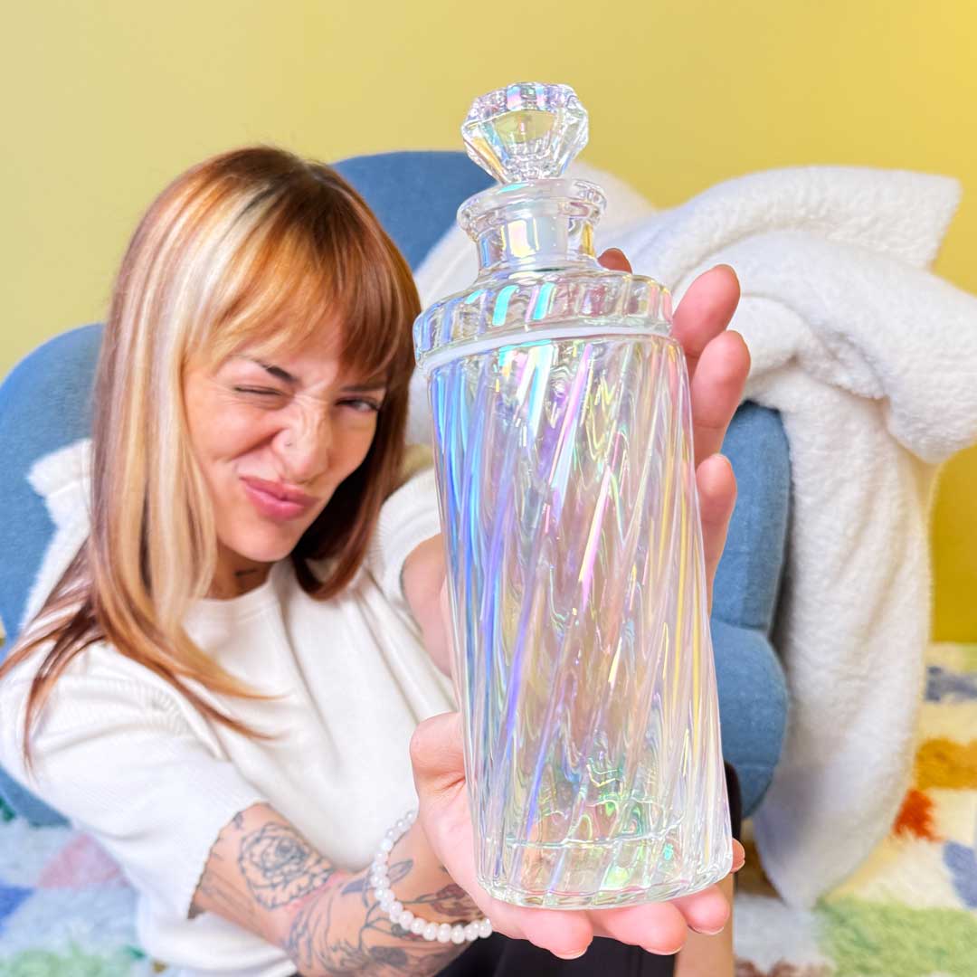 model laying on colorful checkered rug and leaning against a blue chair. Model is holding up an iridescent swirl gravity bong 